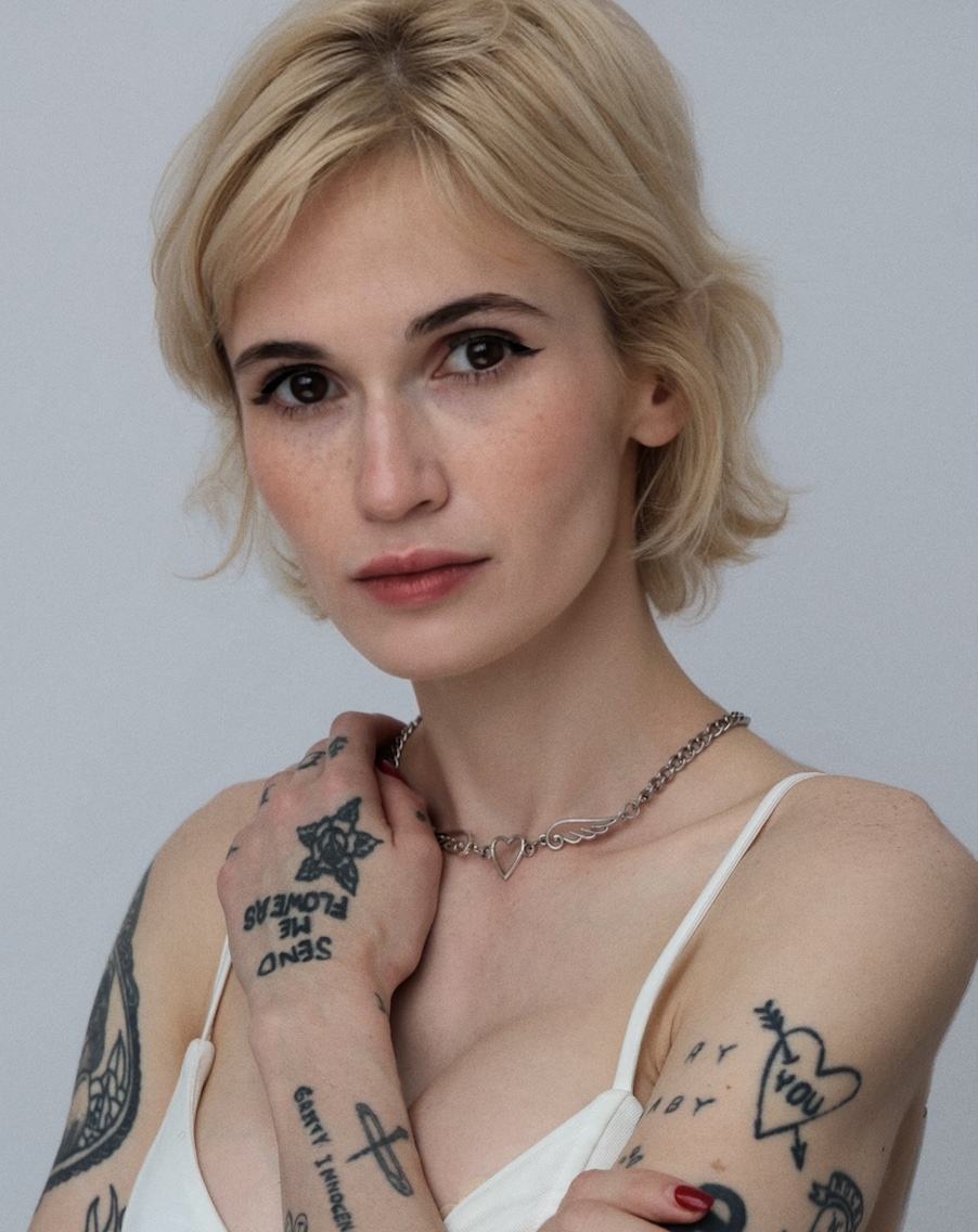 Taylor Drew, hands at her collarbone, tattooed arms and a chain necklace, against a pale grey studio backdrop